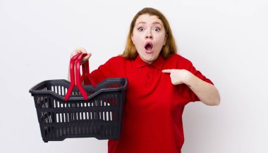 Kızıl saçlı güzel kadın şok olmuş ve ağzı açık, kendini işaret eden bir şekilde şaşırmış görünüyor. boş alışveriş sepeti kavramı