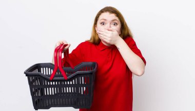 Kızıl saçlı, güzel bir kadın elleri ile ağzını kapatıyor. boş alışveriş sepeti kavramı