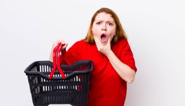 Kızıl saçlı, ağzı ve gözleri açık, eli çenesinde güzel bir kadın. boş alışveriş sepeti kavramı