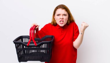 Kızıl saçlı güzel bir kadın sinirli bir ifadeyle agresif bir şekilde bağırıyor. boş alışveriş sepeti kavramı