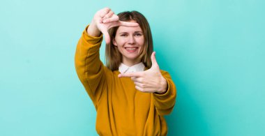 Kızıl saçlı güzel bir kadın mutlu, arkadaş canlısı ve pozitif hissediyor, gülümsüyor ve elleriyle bir portre veya fotoğraf çerçevesi yapıyor.