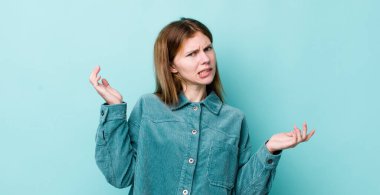 red head pretty woman shrugging with a dumb, crazy, confused, puzzled expression, feeling annoyed and clueless