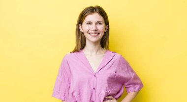 Kızıl saçlı güzel bir kadın mutlu bir şekilde gülümsüyor, eli kalçasında ve kendinden emin, olumlu, gururlu ve arkadaş canlısı bir tavırla.