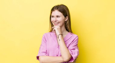 kızıl saçlı güzel bir kadın mutlu, kendinden emin bir ifadeyle gülümsüyor elleri çenesinde, merak ediyor ve yan tarafa bakıyor.