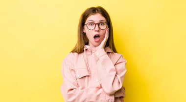 Kızıl saçlı güzel bir kadın şok ve inançsızlıkla ağzı açık, elleri yanağında ve kolları çapraz, sersemlemiş ve şaşırmış hissediyor.