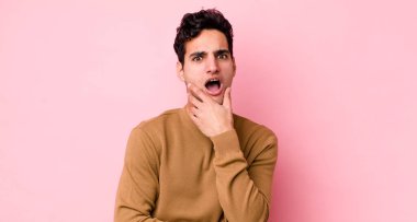 Yakışıklı İspanyol adam şok ve inançsızlık içinde ağzı açık, elleri yanak ve kolları çapraz, sersemlemiş ve şaşkın hissediyor