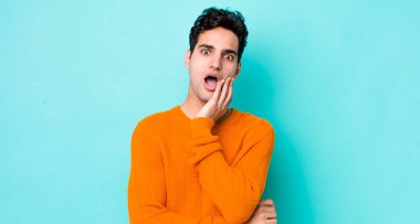 Yakışıklı İspanyol adam şok ve inançsızlık içinde ağzı açık, elleri yanak ve kolları çapraz, sersemlemiş ve şaşkın hissediyor