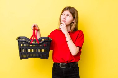 Kızıl saçlı güzel kadın düşünüyor, kuşkulu ve şaşkın hissediyor. Alışveriş sepeti konsepti