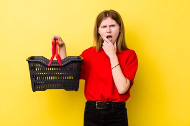 Kızıl saçlı, ağzı ve gözleri açık, eli çenesinde güzel bir kadın. Alışveriş sepeti konsepti