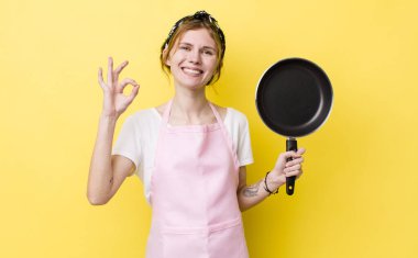 Kızıl saçlı güzel bir kadın kendini mutlu hissediyor. Onayını iyi bir hareketle gösteriyor. kızartma tavası ve ev hanımı kavramı