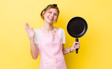 Kızıl saçlı güzel kadın mutlu bir şekilde gülümsüyor, el sallıyor, seni selamlıyor ve selamlıyor. kızartma tavası ve ev hanımı kavramı