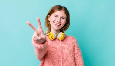 Kızıl saçlı güzel kadın gülümsüyor ve mutlu görünüyor, zafer ya da barış işareti yapıyor. kulaklıklar ve müzik konsepti