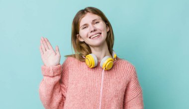 Kızıl saçlı güzel kadın mutlu bir şekilde gülümsüyor, el sallıyor, seni selamlıyor ve selamlıyor. kulaklıklar ve müzik konsepti