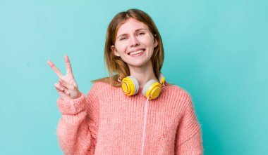 Kızıl saçlı güzel kadın gülümsüyor ve arkadaş canlısı görünüyor, iki numarayı gösteriyor. kulaklıklar ve müzik konsepti