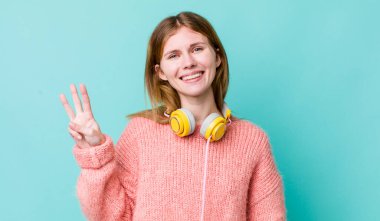 Kızıl saçlı güzel kadın gülümsüyor ve arkadaş canlısı görünüyor, üç numarayı gösteriyor. kulaklıklar ve müzik konsepti
