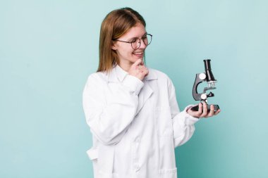 Kızıl saçlı, güzel bir kadın mutlu, kendinden emin, elleri çenesinde gülümsüyor. Mikroskop konseptine sahip bilim adamı