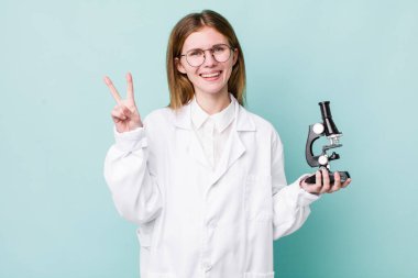 Kızıl saçlı güzel kadın gülümsüyor ve arkadaş canlısı görünüyor, iki numarayı gösteriyor. Mikroskop konseptine sahip bilim adamı