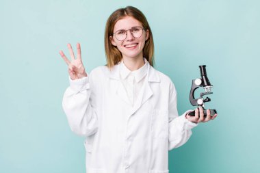 Kızıl saçlı güzel kadın gülümsüyor ve arkadaş canlısı görünüyor, üç numarayı gösteriyor. Mikroskop konseptine sahip bilim adamı