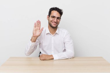 İspanyol yakışıklı adam mutlu bir şekilde gülümsüyor, el sallıyor, seni selamlıyor ve selamlıyor. iş adamı ve masa başı konsepti