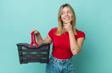 Güzel sarışın kadın gülümsüyor mutlu, kendine güvenen bir yüz ifadesi ve elleri çenesinde. Alışveriş sepeti konsepti