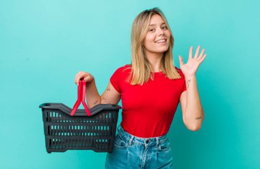 Güzel sarışın kadın mutlu bir şekilde gülümsüyor, el sallıyor, hoş geldin diyor ve seni selamlıyor. Alışveriş sepeti konsepti