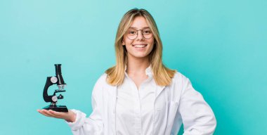 Güzel sarışın kadın mutlu bir şekilde gülümsüyor, eli kalçasında ve kendinden emin. Bilim adamı ve mikroskop kavramı