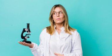 Güzel sarışın kadın omuz silkiyor, kafası karışık ve kararsız. Bilim adamı ve mikroskop kavramı