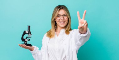Güzel sarışın kadın gülümsüyor ve arkadaş canlısı görünüyor. İki numarayı gösteriyor. Bilim adamı ve mikroskop kavramı