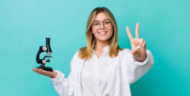 Güzel sarışın kadın gülümsüyor ve mutlu görünüyor, zafer ya da barış işareti yapıyor. Bilim adamı ve mikroskop kavramı