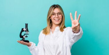 Güzel sarışın kadın gülümsüyor ve arkadaş canlısı görünüyor, üç numarayı gösteriyor. Bilim adamı ve mikroskop kavramı