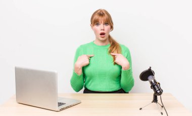 Kızıl saçlı güzel bir kadın mutlu hissediyor ve heyecanla kendini işaret ediyor. dizüstü bilgisayar ve mikrofon kavramı