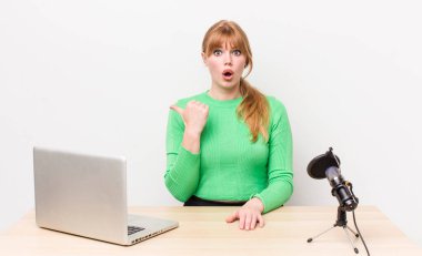 Kızıl saçlı, güzel bir kadın inançsızlıktan şaşkın şaşkın bakıyor. dizüstü bilgisayar ve mikrofon kavramı