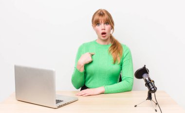 Kızıl saçlı güzel kadın şok olmuş ve ağzı açık, kendini işaret eden bir şekilde şaşırmış görünüyor. dizüstü bilgisayar ve mikrofon kavramı