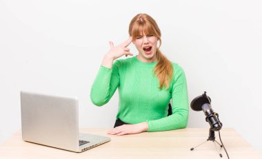 Kızıl saçlı güzel bir kadın mutsuz ve stresli görünüyor, intihar jesti silah işareti yapıyor. dizüstü bilgisayar ve mikrofon kavramı