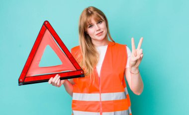 Kızıl saçlı güzel kadın gülümsüyor ve arkadaş canlısı görünüyor, iki numarayı gösteriyor. araba acil üçgeni