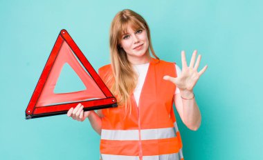 Kızıl saçlı güzel kadın gülümsüyor ve arkadaş canlısı görünüyor, beş numarayı gösteriyor. araba acil üçgeni