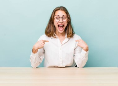 Genç ve güzel bir kadın kendini mutlu hissediyor ve heyecanlı bir şekilde kendini işaret ediyor. masa ve iş konsepti