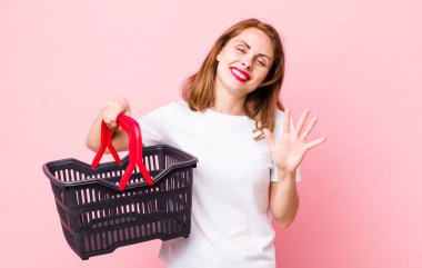 Genç, güzel bir bayan gülümsüyor ve arkadaş canlısı görünüyor, beş numarayı gösteriyor. boş alışveriş sepeti kavramı