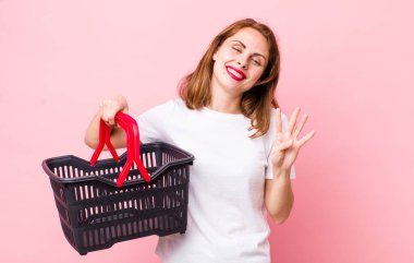 Genç, güzel bir bayan gülümsüyor ve arkadaş canlısı görünüyor, dört numarayı gösteriyor. boş alışveriş sepeti kavramı