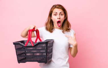 Şok olmuş ve ağzı açık, kendini gösteren genç ve güzel bir kadın. boş alışveriş sepeti kavramı