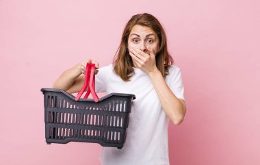 Genç ve güzel bir kadın şok olmuş bir şekilde elleriyle ağzını kapatıyor. boş alışveriş sepeti kavramı