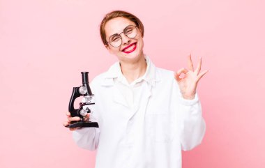 Genç ve güzel bir kadın kendini mutlu hissediyor. Onayını iyi bir hareketle gösteriyor. Mikroskop konseptine sahip bilim adamı
