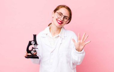 Genç, güzel bir bayan gülümsüyor ve arkadaş canlısı görünüyor, beş numarayı gösteriyor. Mikroskop konseptine sahip bilim adamı