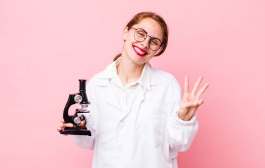 Genç, güzel bir bayan gülümsüyor ve arkadaş canlısı görünüyor, üç numarayı gösteriyor. Mikroskop konseptine sahip bilim adamı