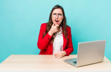 young pretty woman  with mouth and eyes wide open and hand on chin. business desk concept