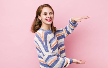 Genç, güzel bir kadın gülümsüyor, mutlu, pozitif ve tatmin olmuş hissediyor, fotokopi alanında nesne veya konsept tutuyor veya gösteriyor