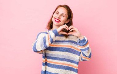 Genç ve güzel bir kadın gülümsüyor ve mutlu, sevimli, romantik ve aşık, iki eliyle kalbi şekillendiriyor