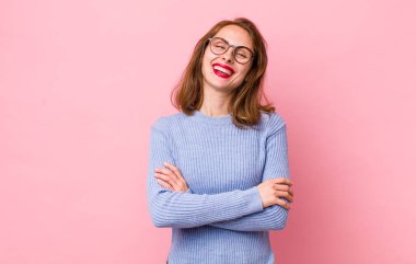 Genç ve güzel bir kadın kollarını kavuşturup mutlu bir şekilde gülüyor, rahat, pozitif ve tatmin edici bir pozla.