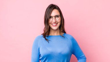 Genç ve güzel bir kadın mutlu bir şekilde gülümsüyor, eli kalçasında ve kendinden emin, pozitif, gururlu ve arkadaş canlısı.