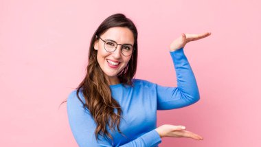 Genç, güzel bir kadın gülümsüyor, mutlu, pozitif ve tatmin olmuş hissediyor, fotokopi alanında nesne veya konsept tutuyor veya gösteriyor